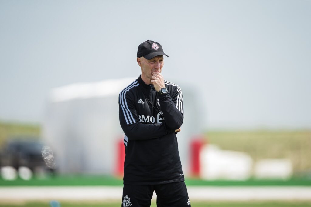 Một huấn luyện viên mặc bộ đồ thể thao màu đen của Toronto FC, đang trầm ngâm quan sát các cầu thủ trong một buổi tập luyện.