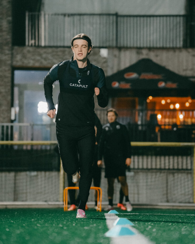 Giocatore di calcio che si allena su un campo illuminato, indossando la tecnologia di monitoraggio Catapult per monitorare il movimento e la fatica.