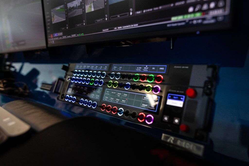 Tampilan close-up dari panel komunikasi dan kontrol yang diterangi yang digunakan pada FIA Race Control, menunjukkan tombol berlabel, sakelar, dan antarmuka layar sentuh yang disematkan.