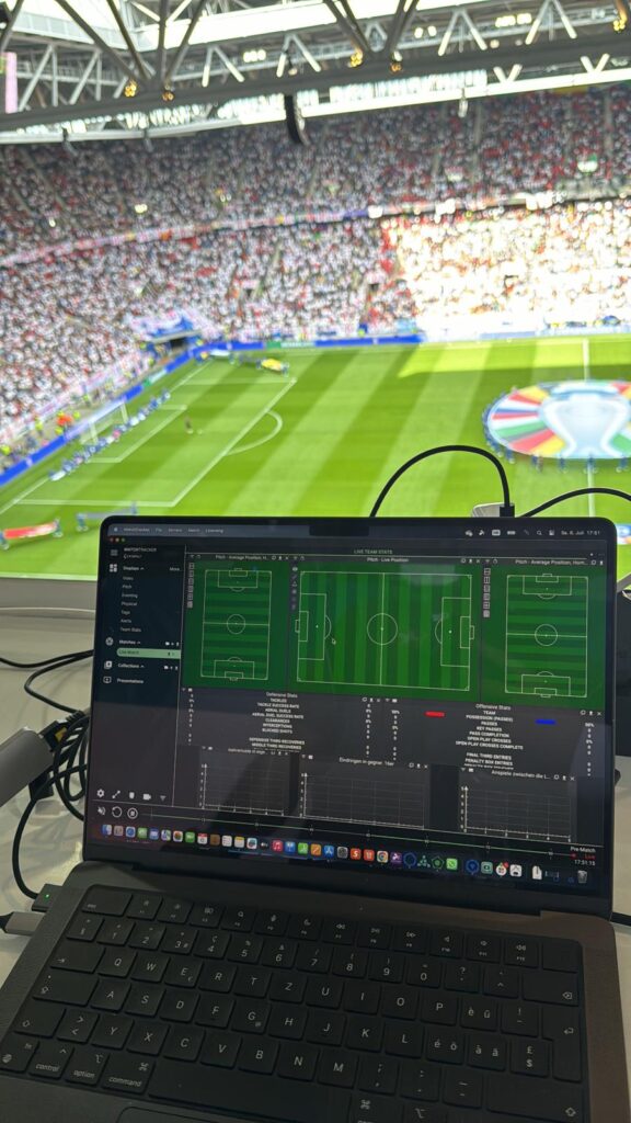 Laptop yang menampilkan antarmuka Catapult MatchTracker selama pertandingan sepak bola langsung di stadion yang penuh sesak.