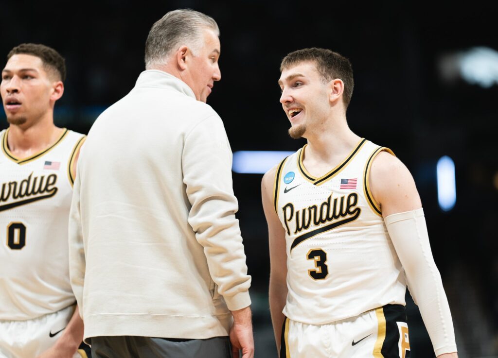 Purdue Boilermakers kosárlabdaedző bézs színű melegítőben beszélget egy mosolygó játékossal, aki fehér és aranyszínű, 3-as számmal ellátott egyenruhát visel a pálya szélén.