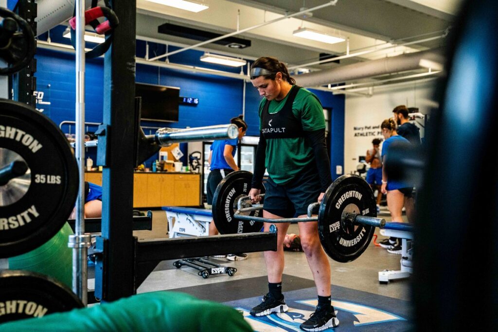 Giocatrice di calcio femminile della Creighton che esegue deadlift in palestra, indossando i giubbotti di monitoraggio Catapult per monitorare i dati dell'allenamento della forza.